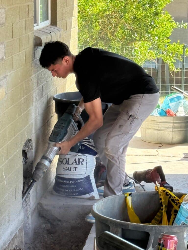 Plumber kneeling and clearing an outdoor drain line or sewer connection on the side of a building.
