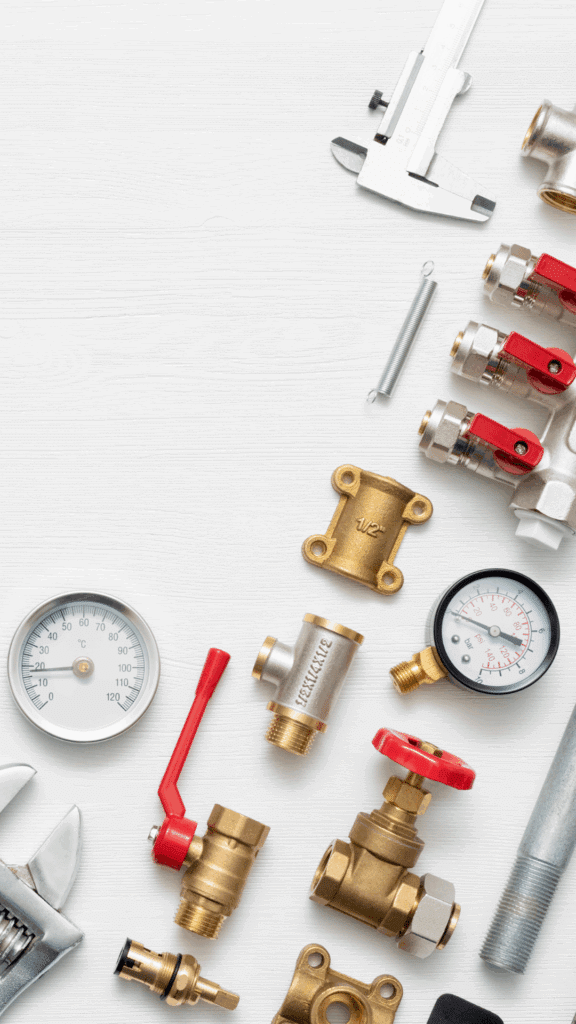 Overhead view of various plumbing tools, brass fittings, red-handled valves, and pressure gauges on a white wooden background.