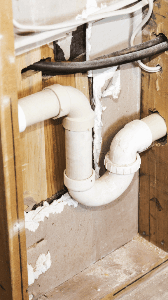 Close-up of white PVC drain pipes installed inside a wall cavity with the drywall removed, showing a P-trap or drainage connection.